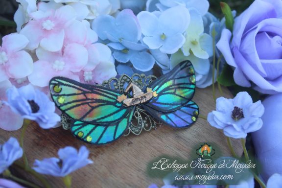 Barrette cheveux ailes de papillon vert turquoise avec filigrane dore, vue rapprochee sur fond de fleurs