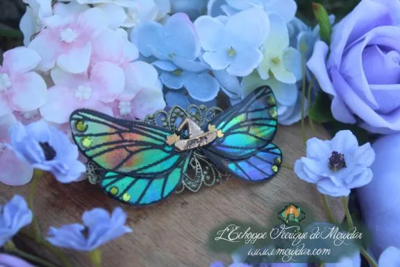 Barrette cheveux ailes de papillon vert turquoise avec filigrane dore, vue rapprochee sur fond de fleurs