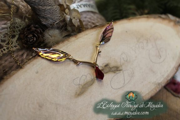 Collier pendentif branche doree avec deux ailes brunes dorees et perle rouge, gros plan sur bois