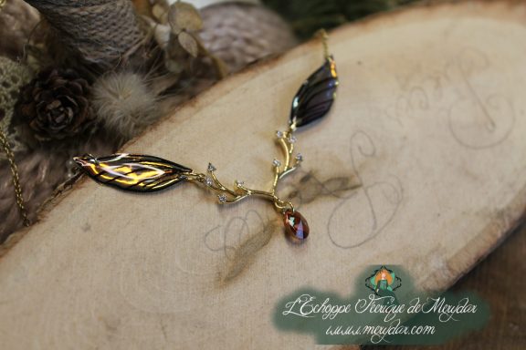 Collier pendentif branche doree avec deux ailes noires vertes et perle rouge, gros plan sur bois