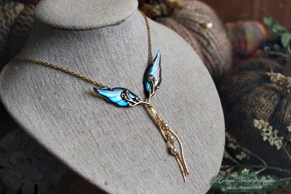 Collier pendentif branche doree avec aile bleue fine, presente sur buste porte-bijoux