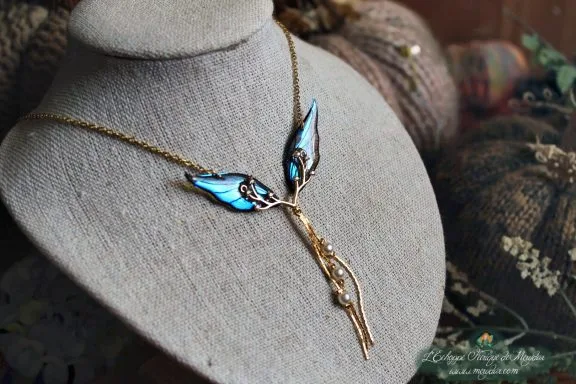 Collier pendentif branche doree avec aile bleue fine, presente sur buste porte-bijoux