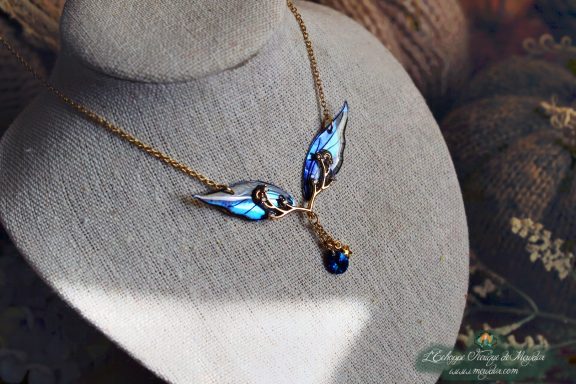 Collier pendentif branche doree avec deux petites ailes bleues et perle bleue, presente sur buste