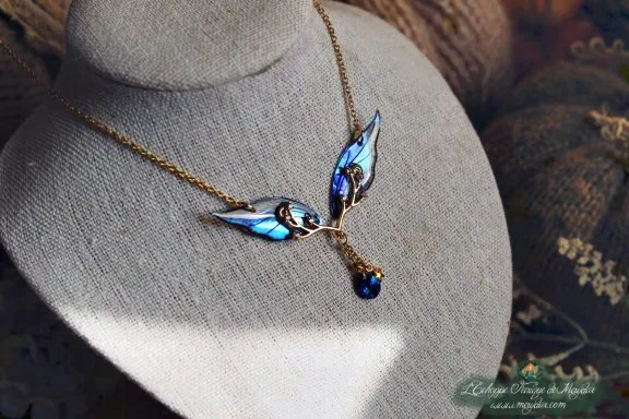 Collier pendentif branche doree avec deux petites ailes bleues et perle bleue, presente sur buste