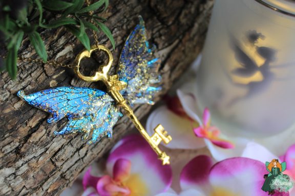 Pendentif cle ailee bleue pailetee doree, presente sur fleurs