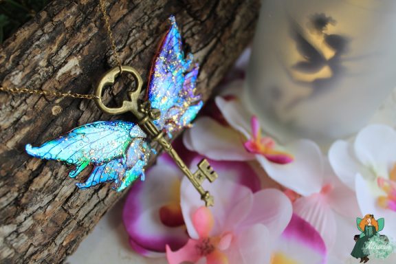 Pendentif cle ailee bleue violette doree, presente sur fleurs